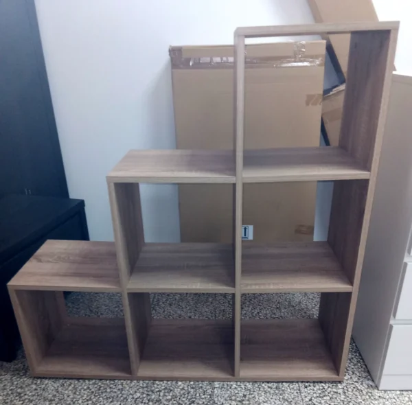 Stufen-Regal in Eiche Nachbildung. Unterste Ebene mit 3 quadratischen Abteilen, mittlere Ebene mit 2 quadratischen Abteilen, oberste Ebene mit einem quadratischen Abteil. Die rechte Regalseite ist über die ganze Höhe gerade, nach links fallen die "Stufen" ab.